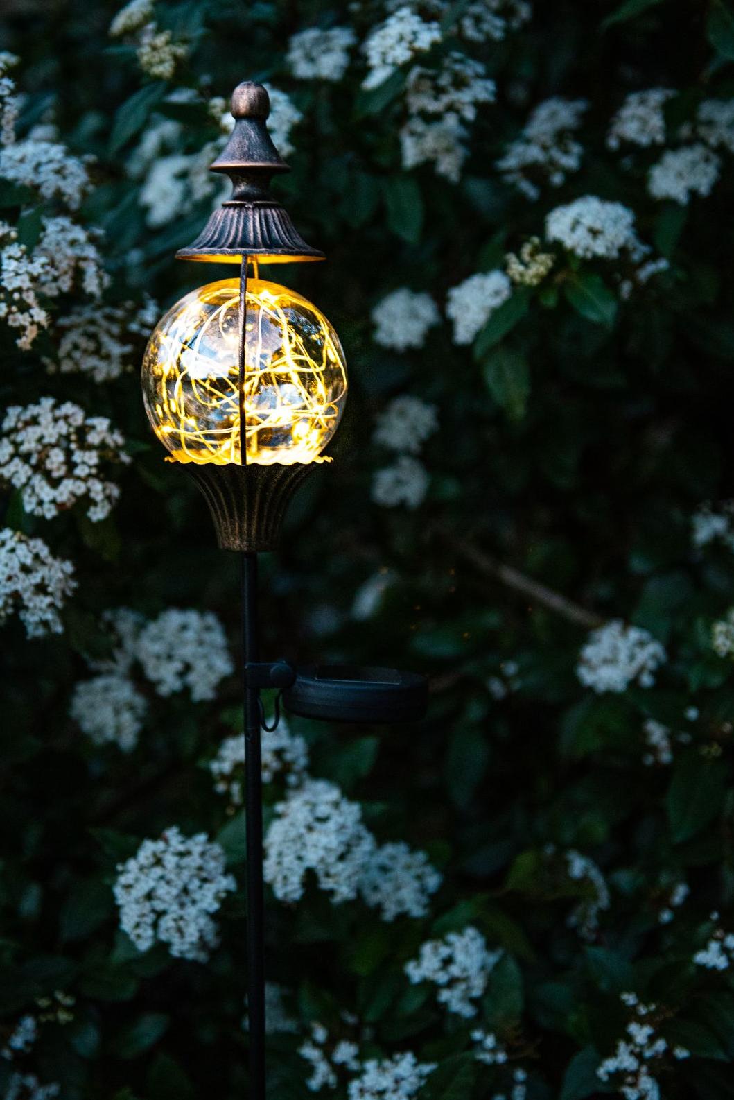 Dekorative Solarlampe im Anthik-Stil auf einem Pfahl mit einer Kugel, die einen Glühfaden mit kleinen Lichtern enthält, der von einer rostfarbenen spitzen Metallkappe umgeben ist Garden ID